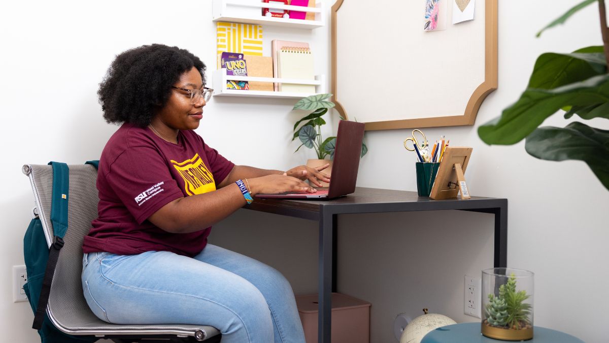 A person sitting at a desk working on a laptop, with a backpack, plants, and office supplies around them.